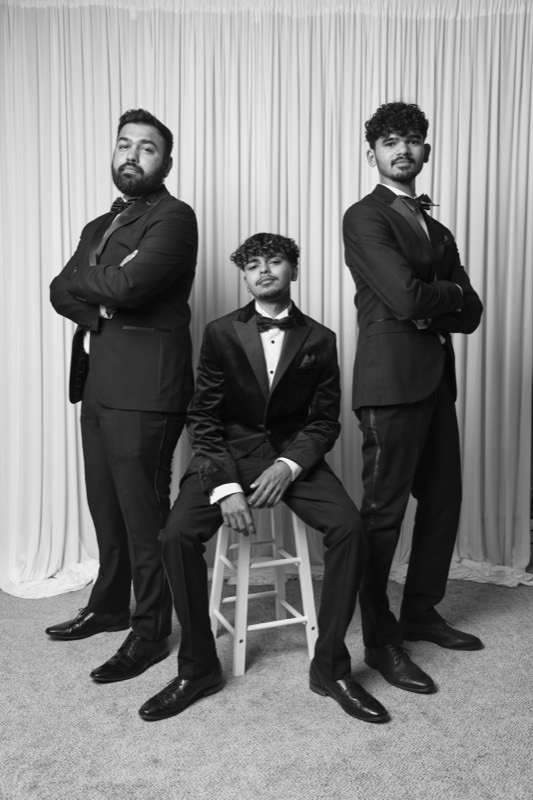 Three young men in black tie pose arms crossed, one seated on a stool