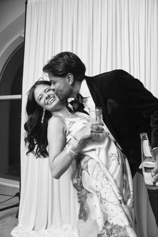 A man in a velvet jacket kisses a woman on the cheek as she leans back laughing
